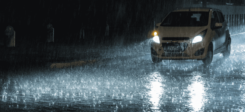 Car driving safely on a wet road during the monsoon season with headlights on, demonstrating safe monsoon driving conditions.